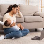 Exhausted Mother Holding Crying Baby And Touching Head Suffering From Headache Sitting At Laptop On Floor At Home. Motherhood Burnout Concept. Tired Mother Posing With Shouting Child