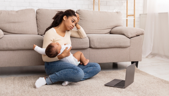 Exhausted Mother Holding Crying Baby And Touching Head Suffering From Headache Sitting At Laptop On Floor At Home. Motherhood Burnout Concept. Tired Mother Posing With Shouting Child